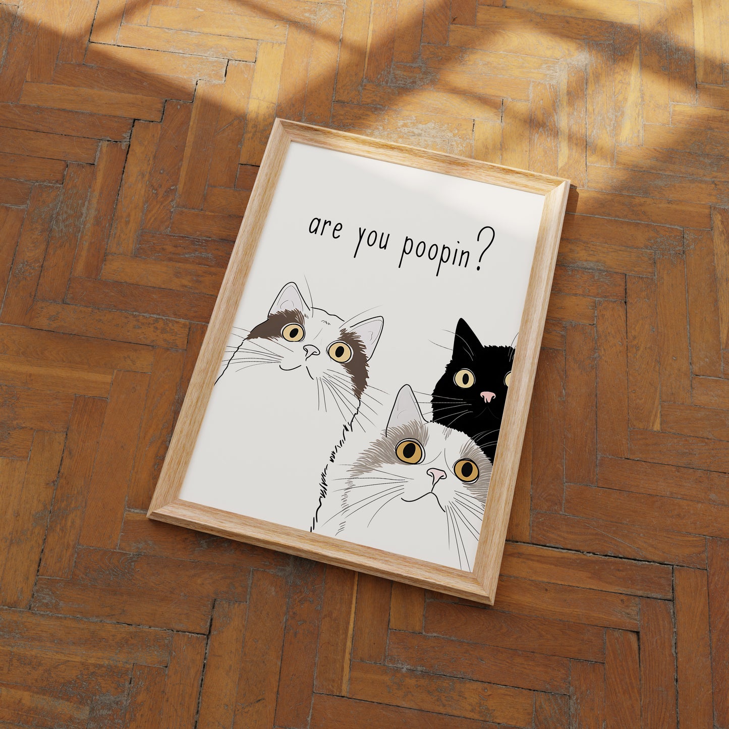 A framed illustration of three curious cats with the caption "are you poopin?" on a wooden floor.