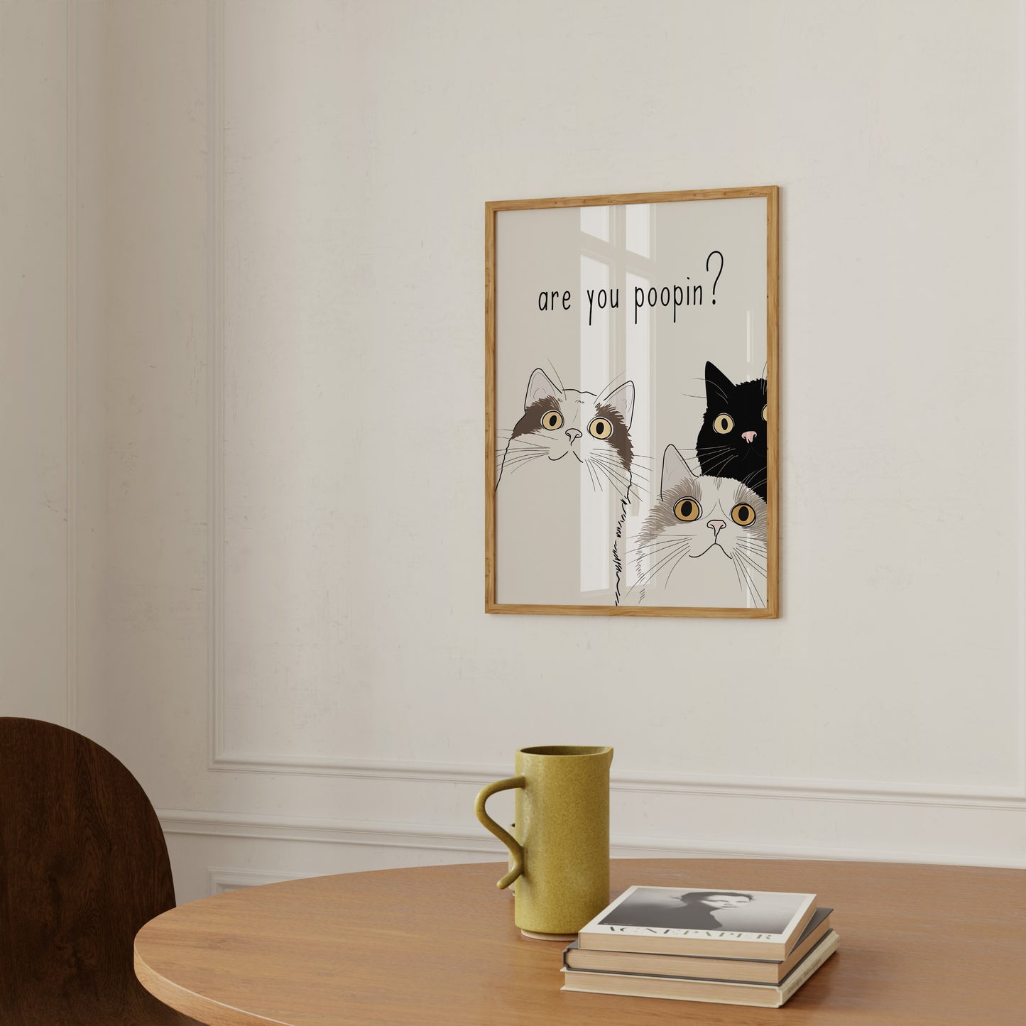 A humorous framed picture on a wall with cats asking "are you poopin?" next to a yellow mug and a book on a table.