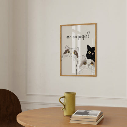 A humorous framed picture on a wall with cats asking "are you poopin?" next to a yellow mug and a book on a table.