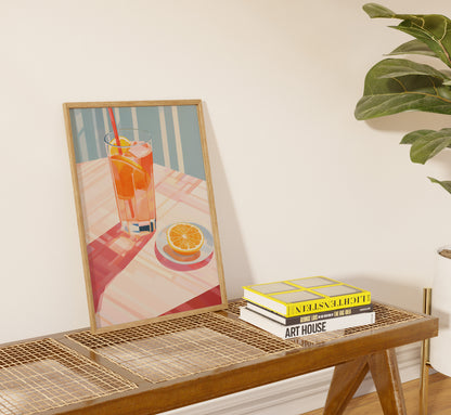 A framed painting of an orange drink beside books on a modern table with a plant.