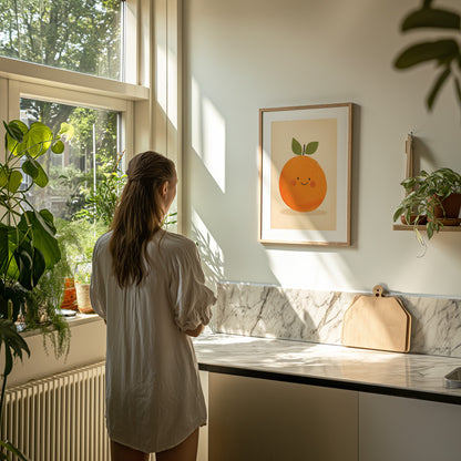 A woman in a white shirt standing in a sunlit kitchen looking at a framed picture of an orange on the wall.
