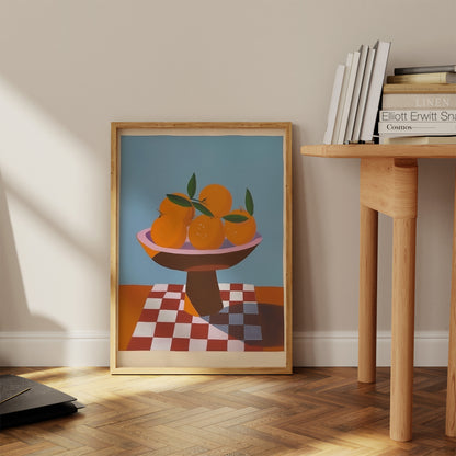 A framed painting of oranges on a table beside books in a sunny room.