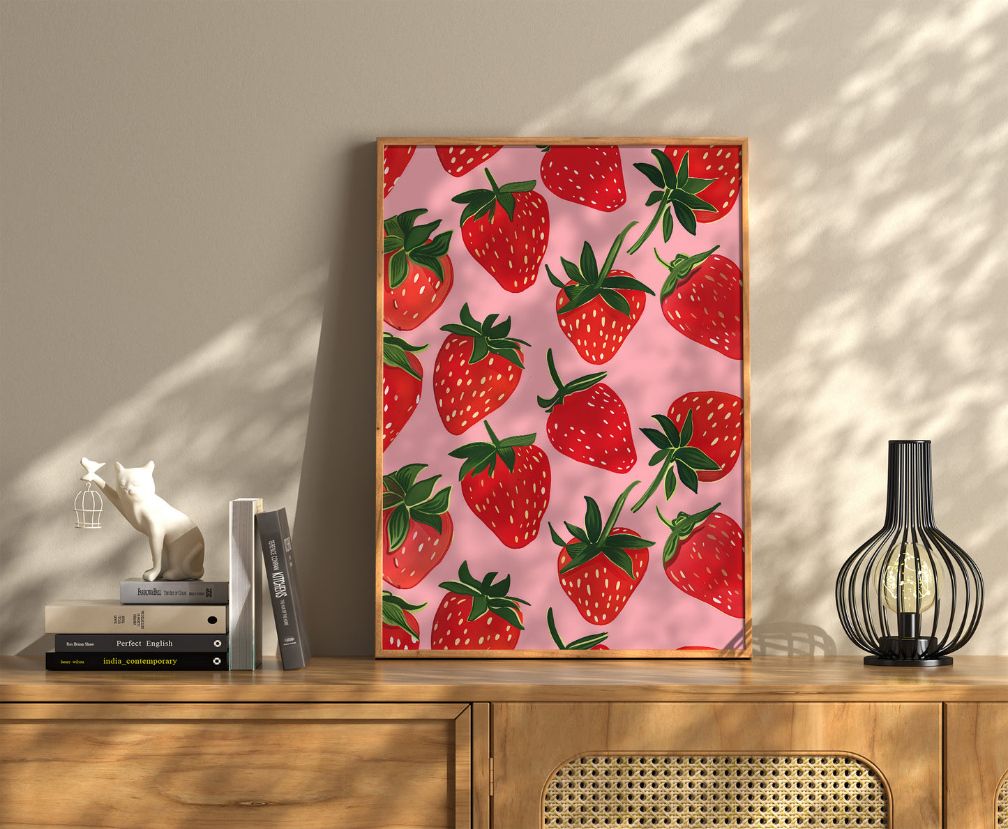 A framed poster of strawberries on a pink background, hanging above a wooden sideboard.