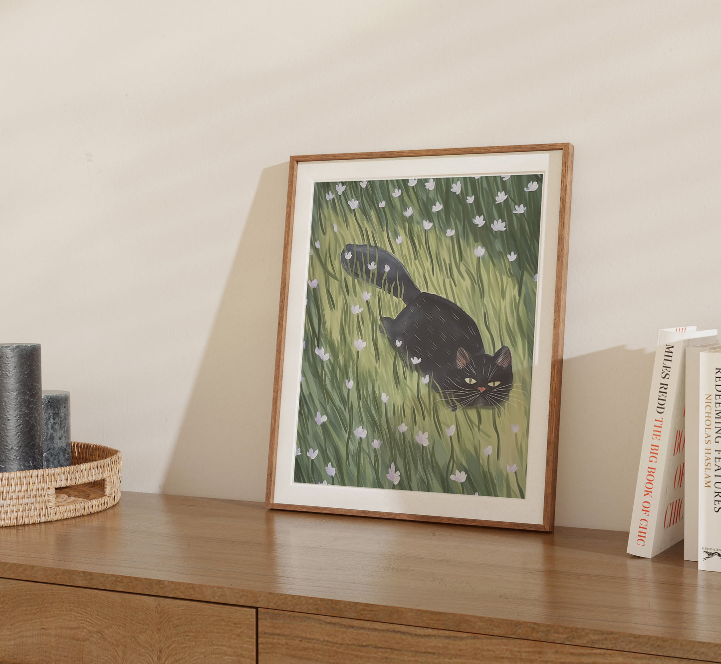 Framed illustration of a black cat in tall grass on a wooden shelf beside books.