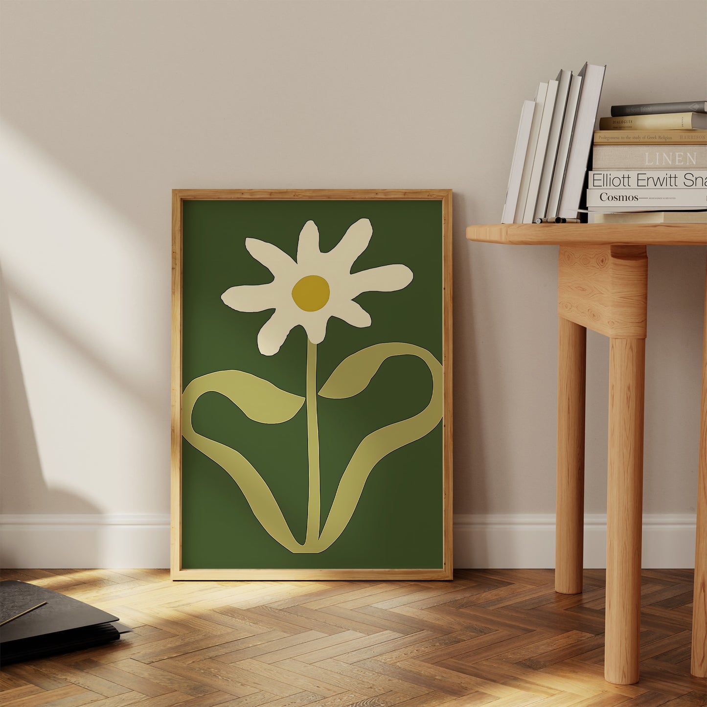 A framed painting of a simplistic white flower on a green background, leaning against a wall next to a wooden table.
