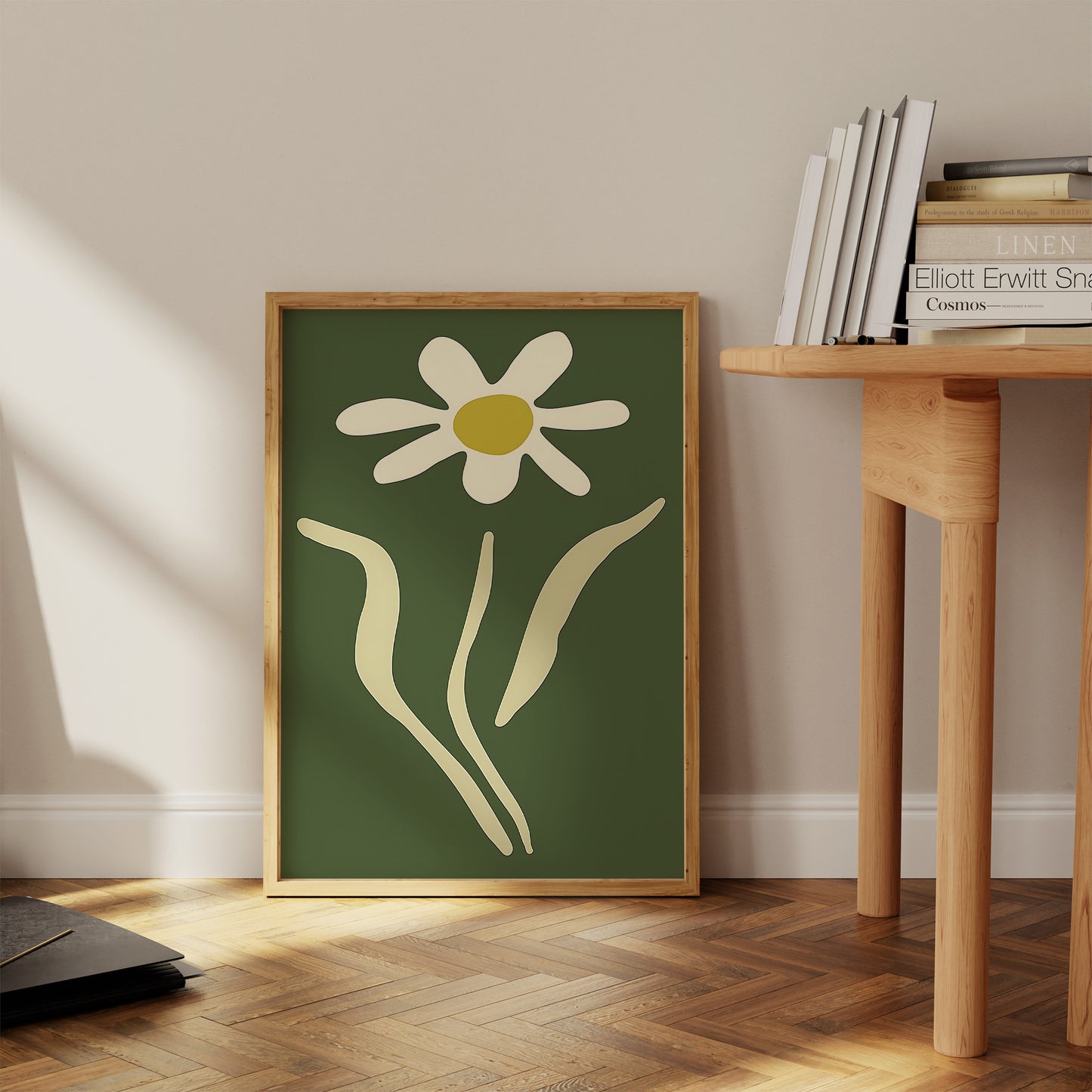 A framed abstract painting of a white flower against a green background leaning against a wall next to a wooden table.