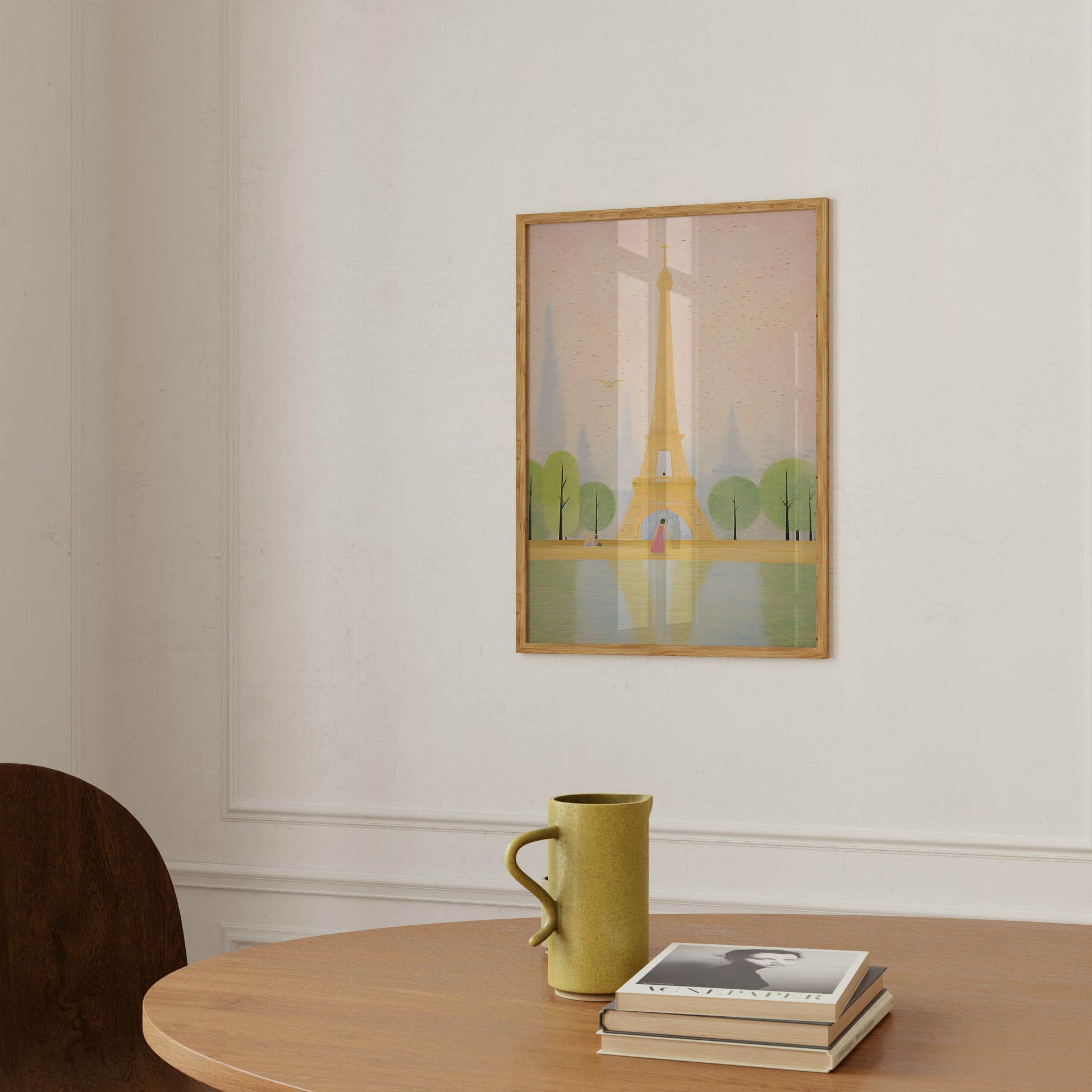 A framed illustration of the Eiffel Tower hangs on a wall above a table with a mug and books.
