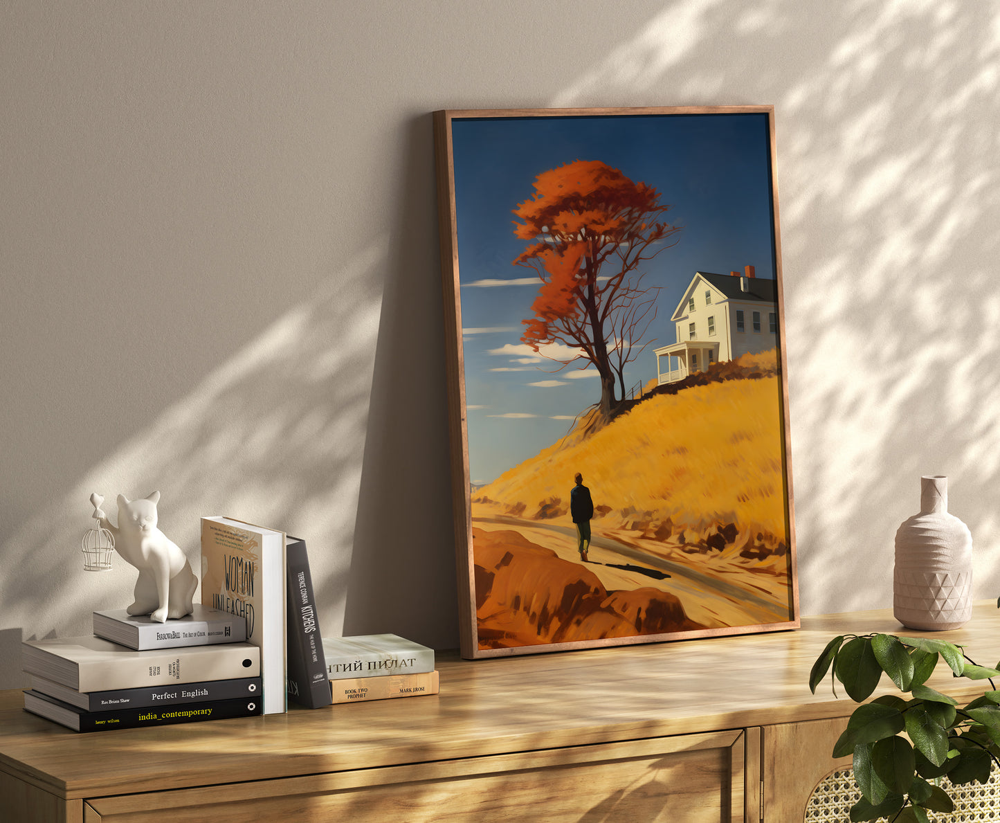 A framed painting of a person walking towards a house with autumn trees, placed in a room with books and decor.