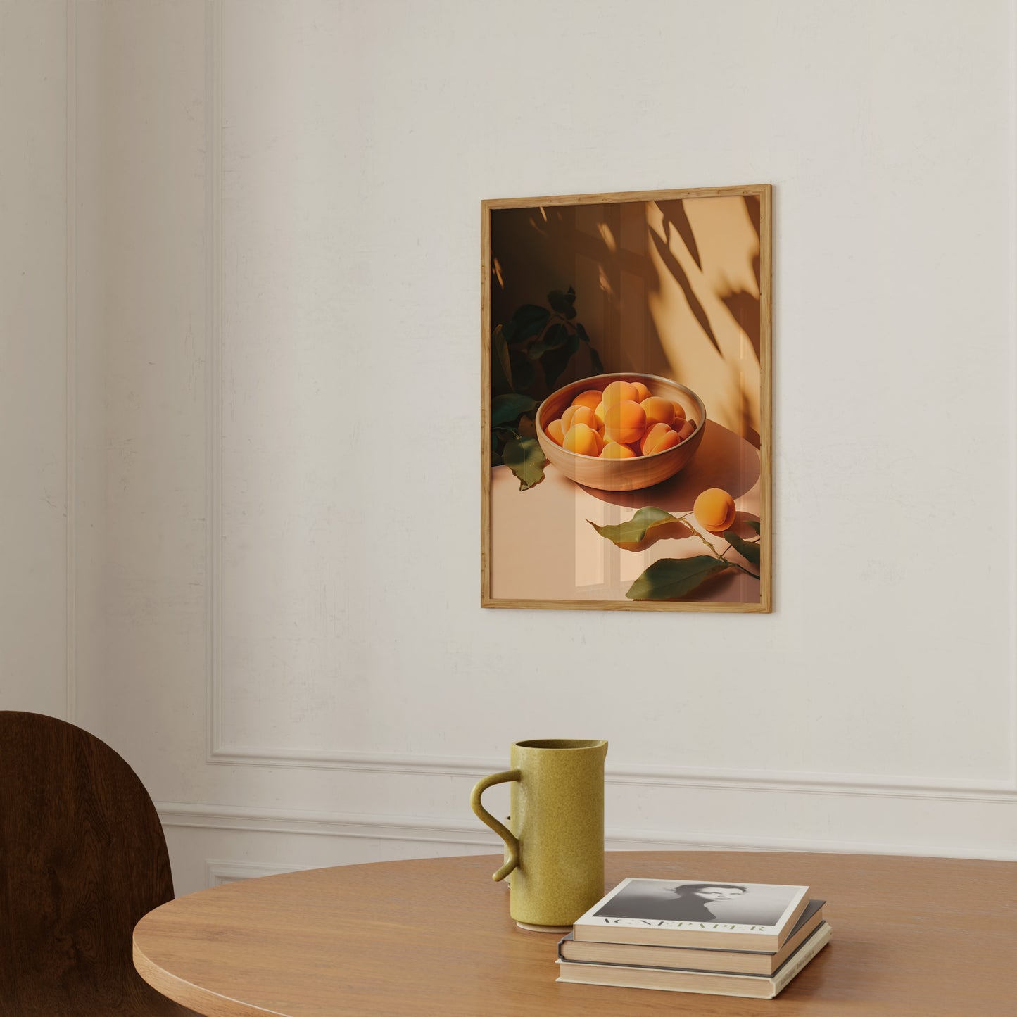 A framed painting of a bowl of oranges on a wall above a table with a mug and books.