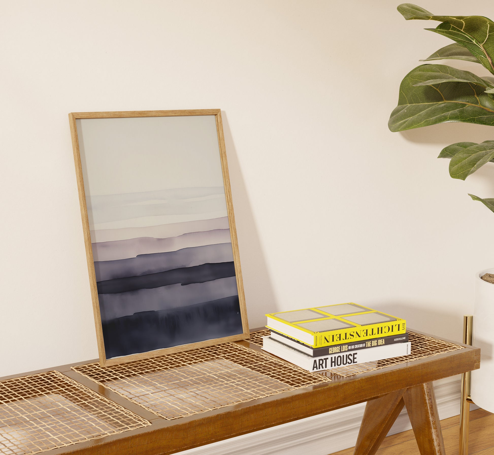 A framed abstract painting leaning against a wall above a wicker bench with books.