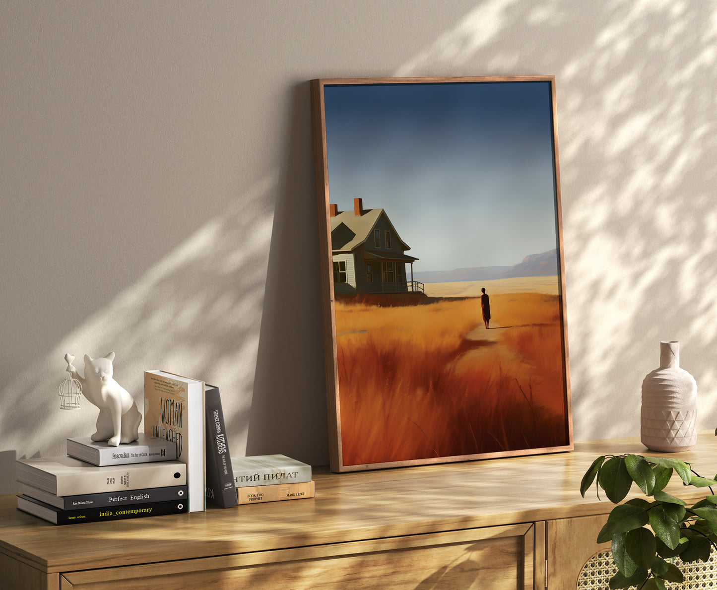 A framed painting of a rural landscape at sunset leaning against a wall beside a shelf with books and decor.