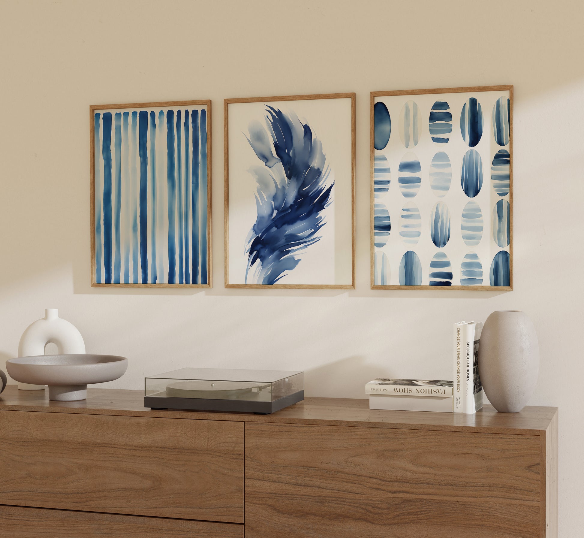 Three abstract paintings in blue tones on a beige wall above a wooden sideboard with decorative items.