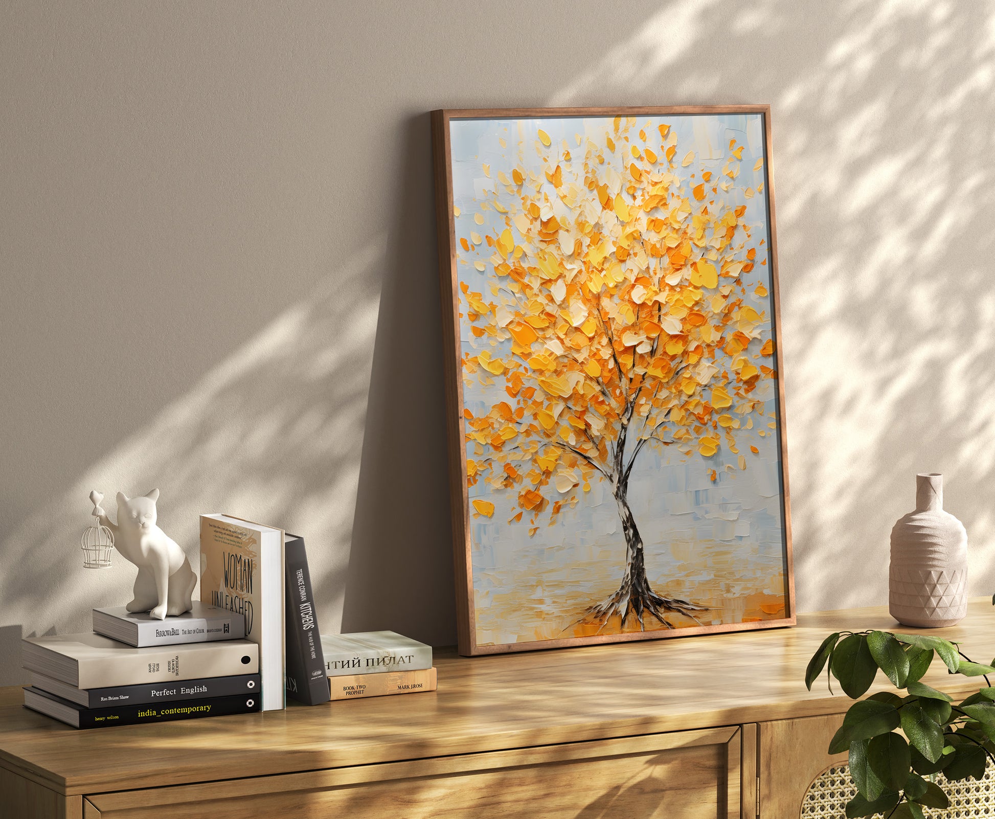 A framed autumn tree painting leaning against a wall beside books and decorative items on a sideboard.