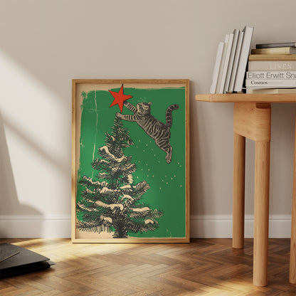 A framed poster of a cat jumping toward a star atop a Christmas tree, leaning against a wall.