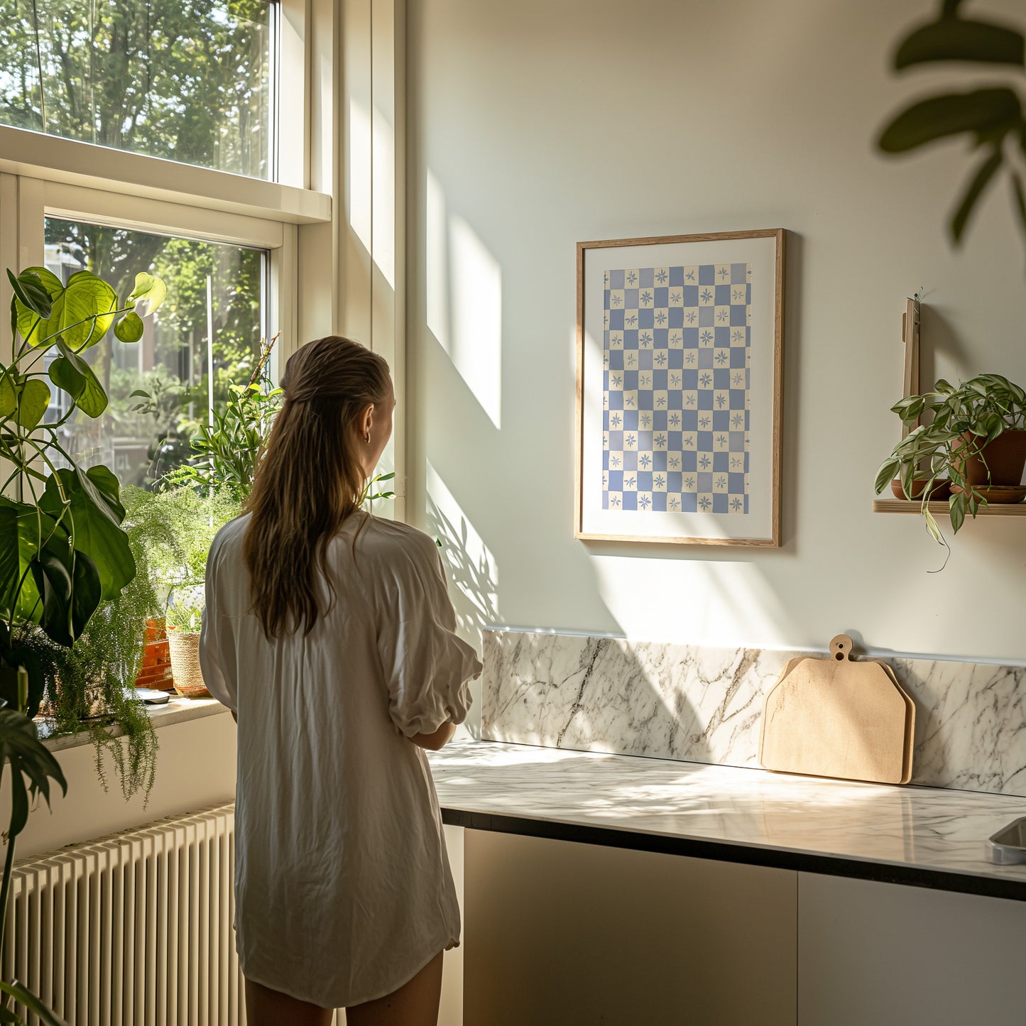Woman in a white shirt looking out of a sunny kitchen window with plants on the sill.