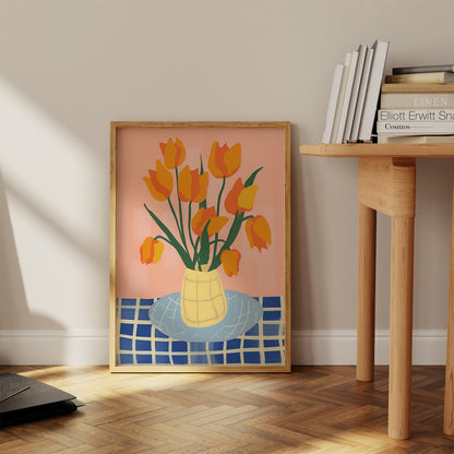 A framed painting of yellow tulips in a vase, beside a wooden table with books, in a room with sunlight.
