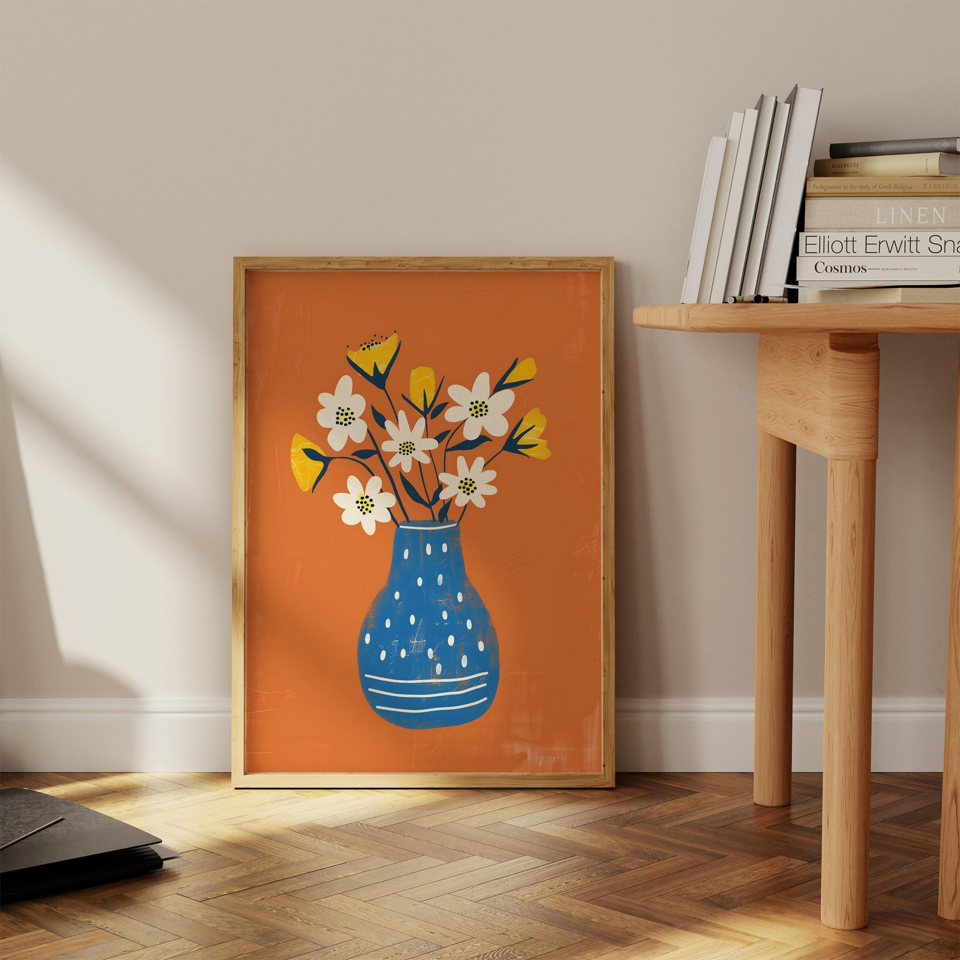 A framed illustration of a blue vase with flowers leaning against a wall beside a wooden table.