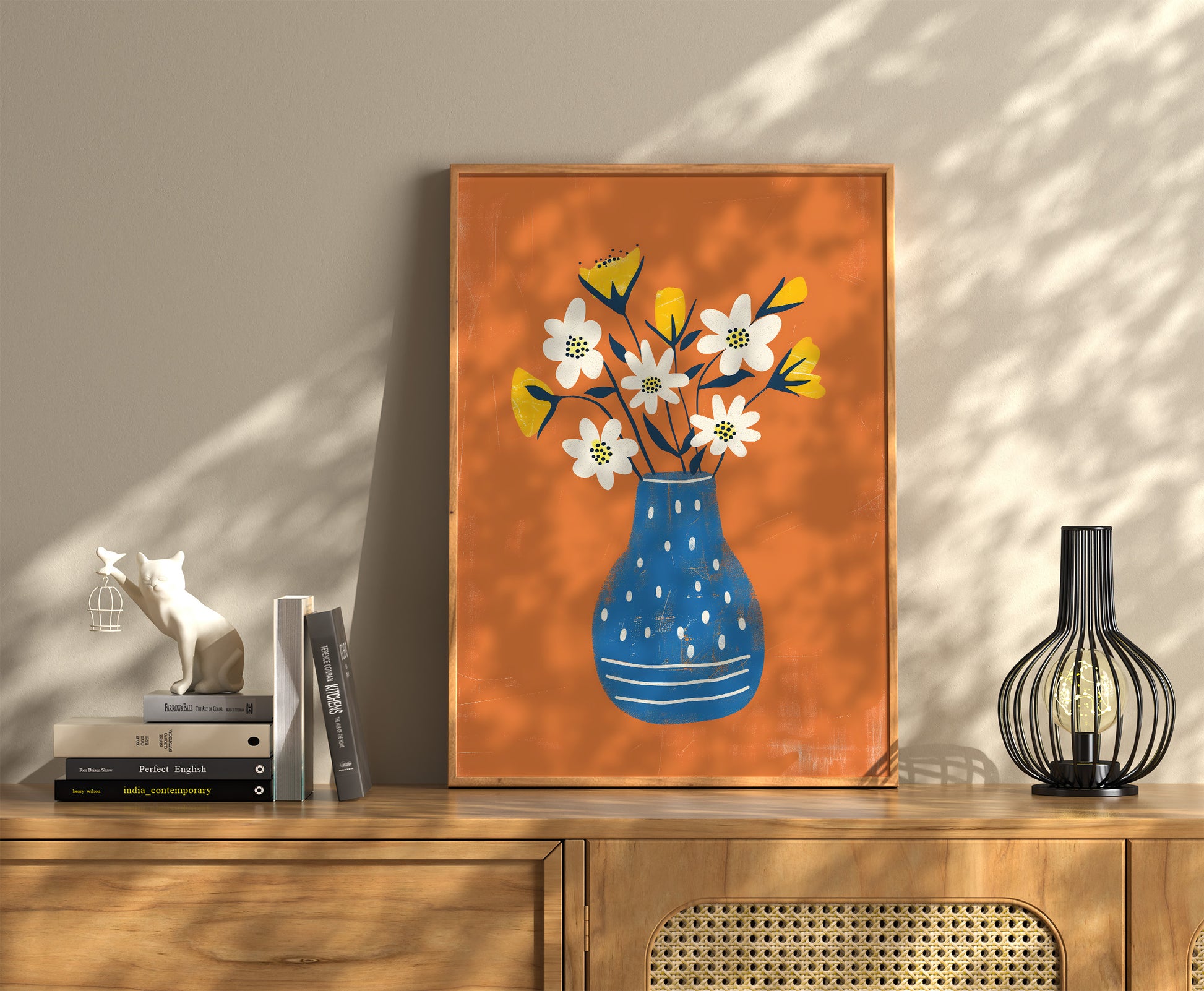 A framed illustration of flowers in a blue vase on a wooden sideboard with decor items.