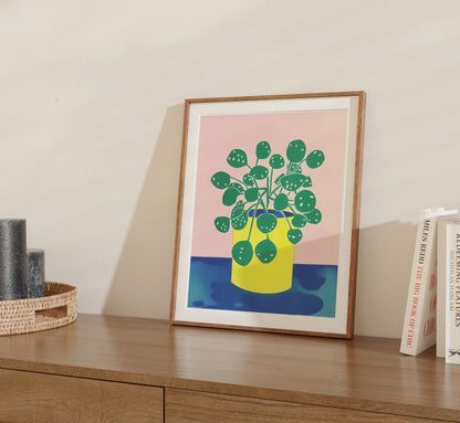 A framed illustration of a potted plant on a shelf with books.
