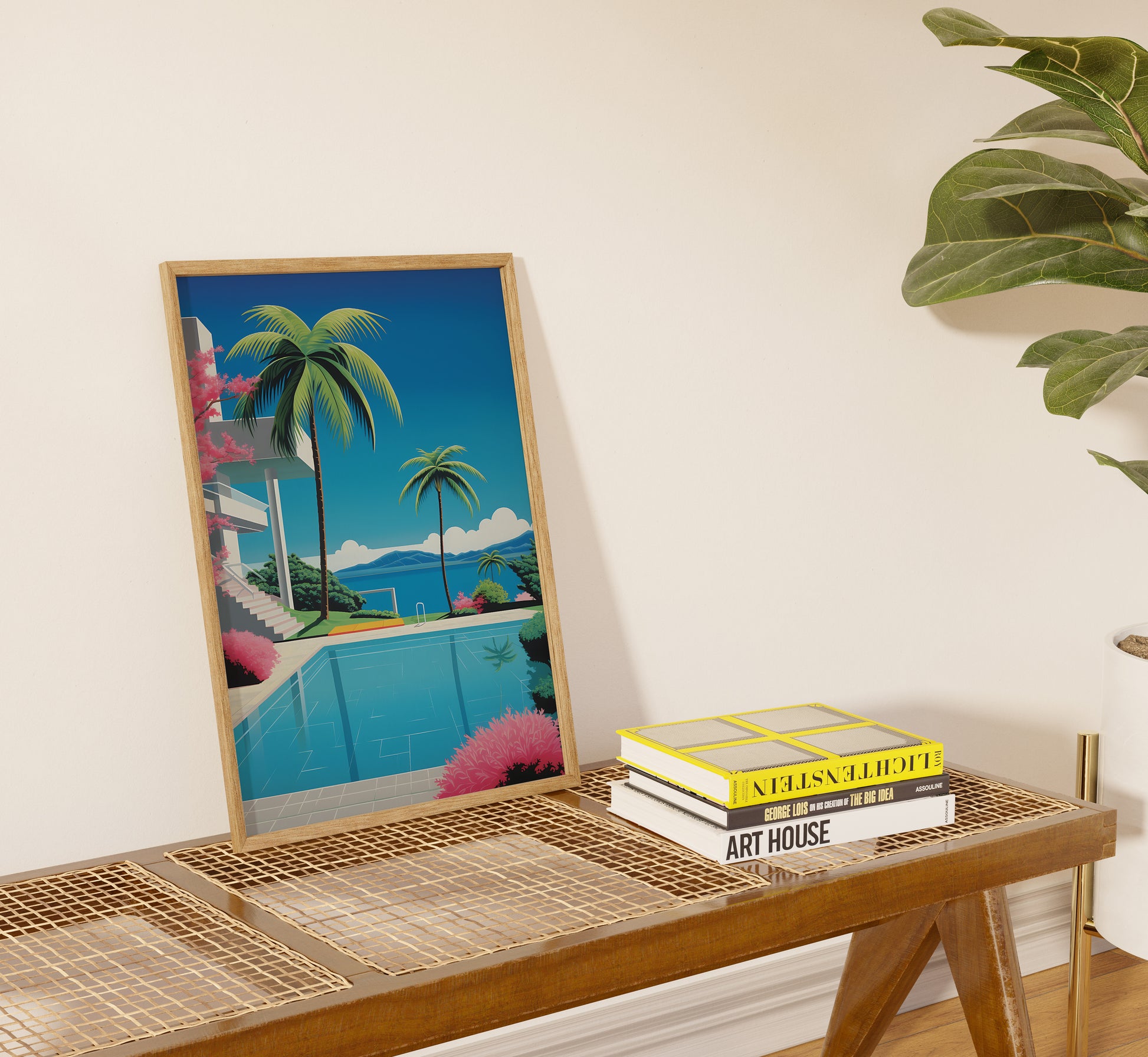 A framed tropical landscape poster next to books on a woven table, with a plant nearby.