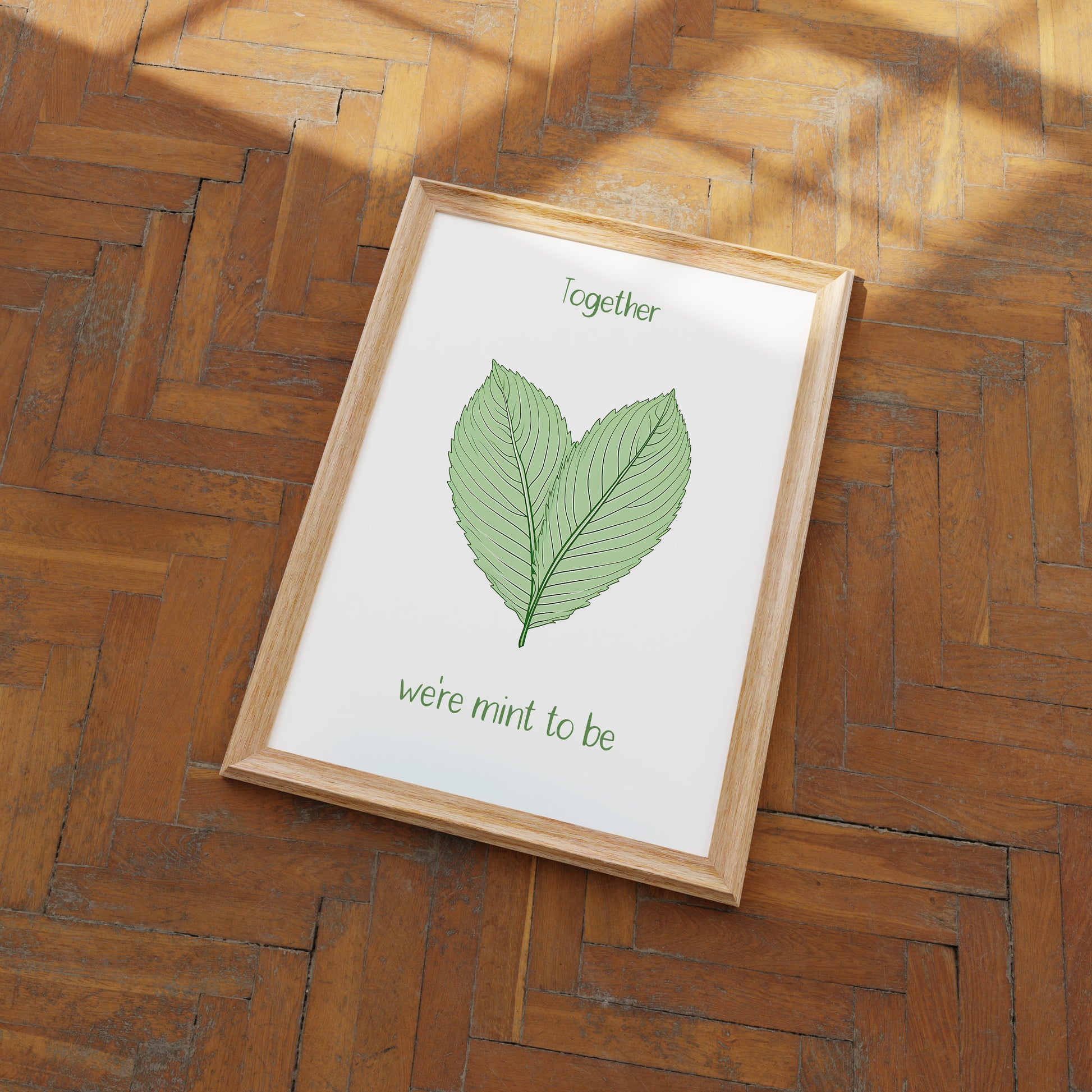 A framed picture with two leaves and the words "Together we're mint to be" on a wooden floor.