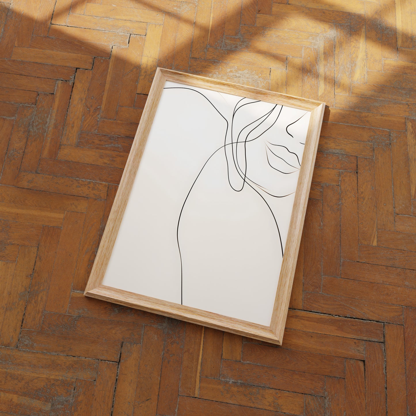 A minimalist line art portrait in a wooden frame lying on a herringbone pattern wood floor.
